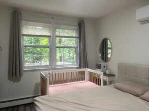 Un dormitorio con una cuna, una mesita de noche con lámpara, un espejo y una ventana con cortinas.