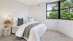 Um quarto com cama, mesa de cabeceira, abajur e janela com vista para árvores.