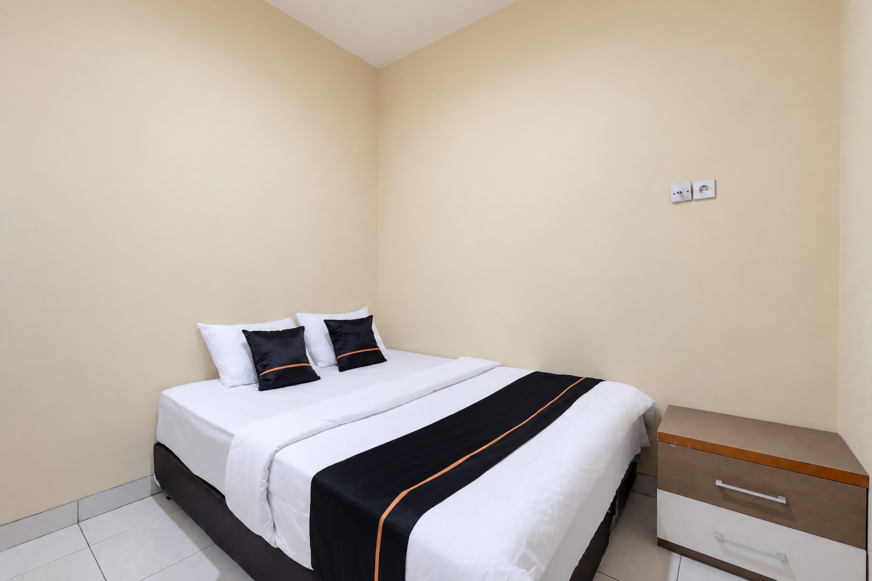 Una cama individual con ropa de cama blanca y dos almohadas negras, una mesita de noche de madera con cajones y un piso de baldosas.