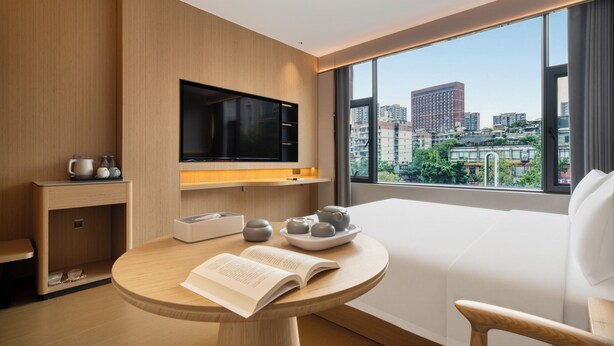 Una habitación de hotel moderna con un ventanal grande, un televisor de pantalla plana, una mesa redonda con un libro y teteras, y un sofá blanco.