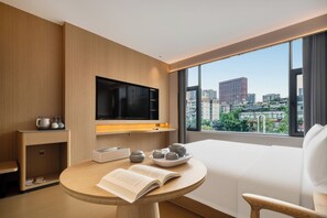 Ein modernes Hotelzimmer mit einem großen Fenster, einem Flachbildfernseher, einem runden Tisch mit einem Buch und Teekannen sowie einem weißen Sofa.