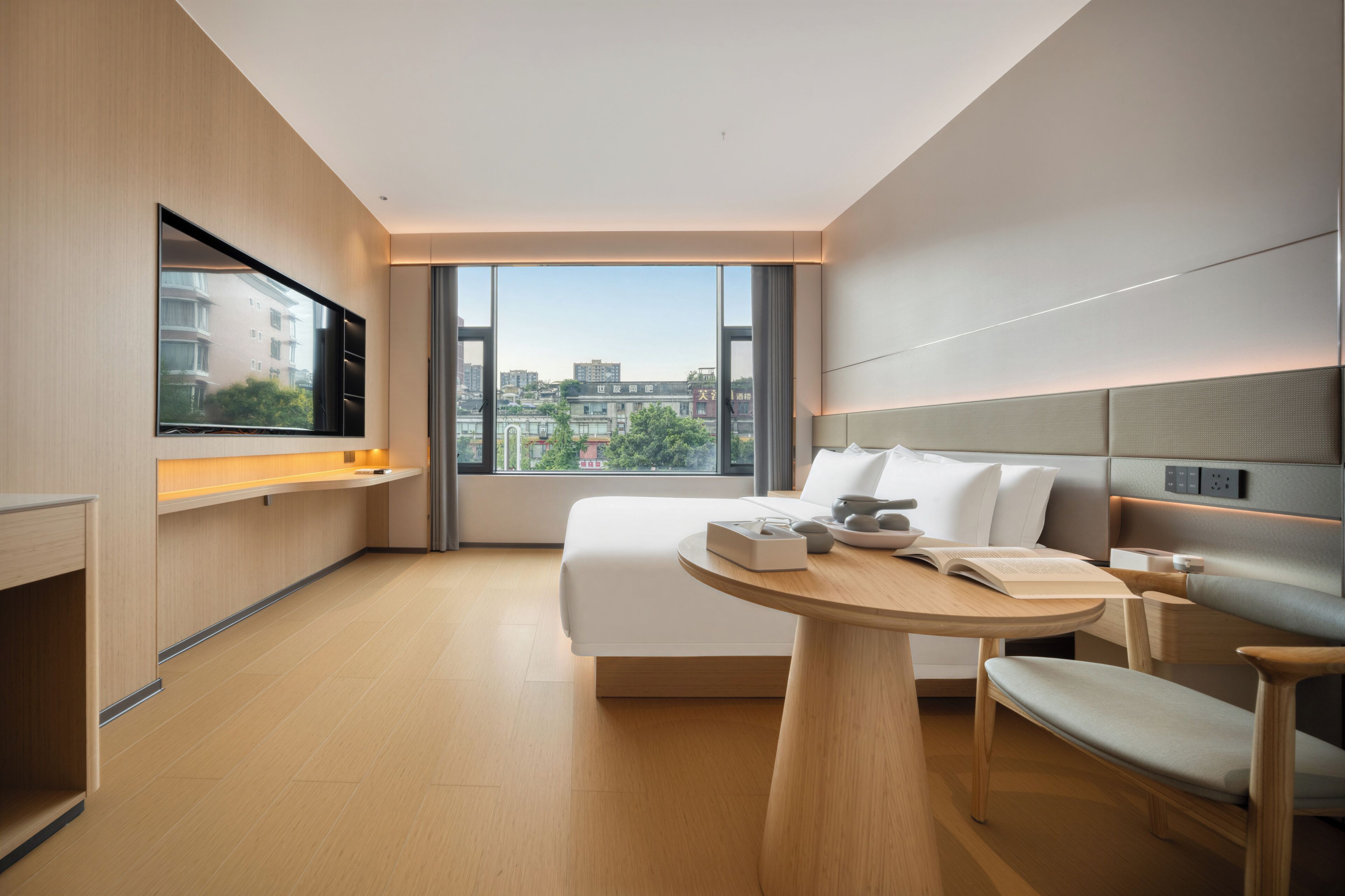 A modern hotel room with a bed, bedside table, chair, and a large window offering a city view.