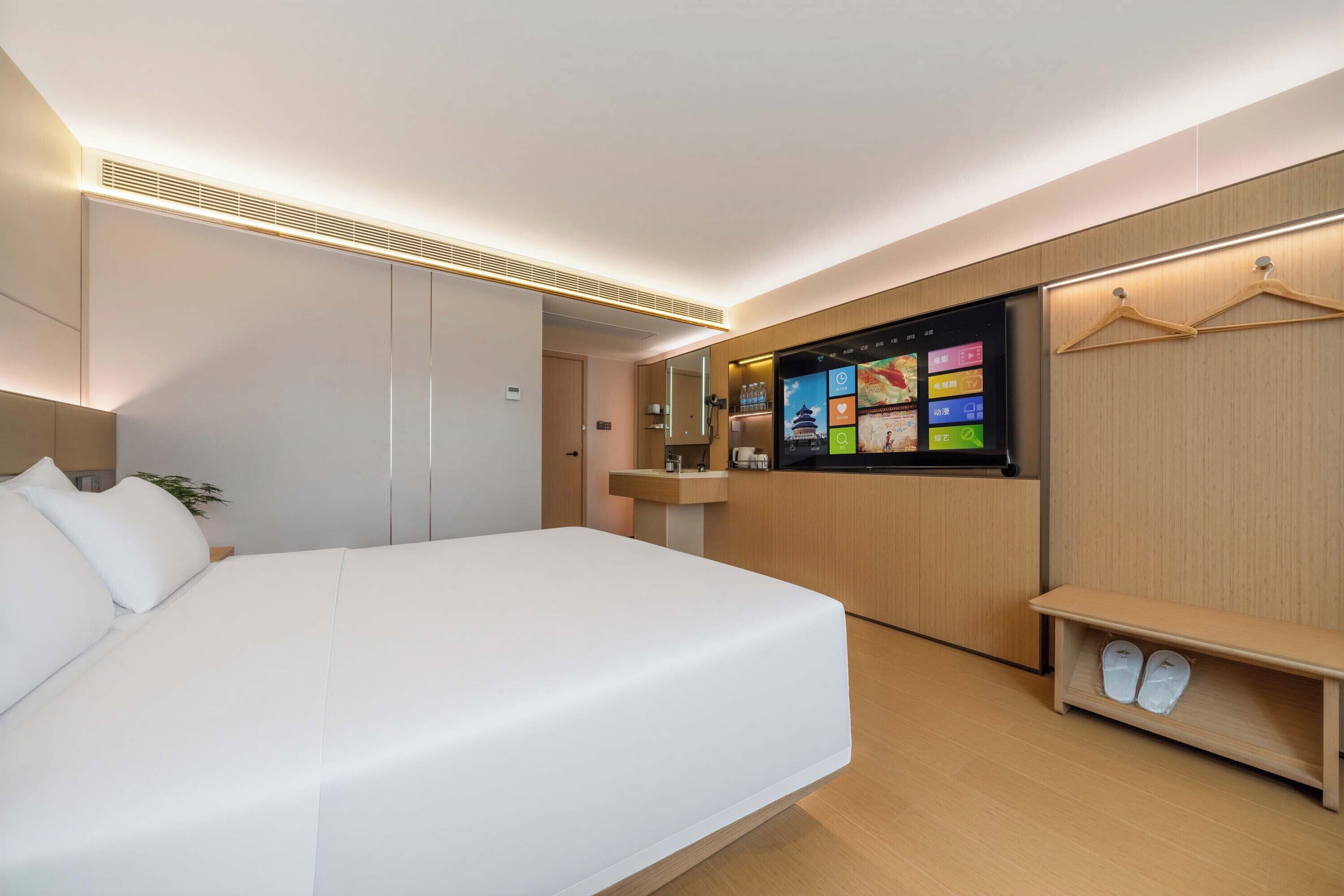 Una camera d'albergo moderna con un letto grande, una TV fissata al muro, un armadio in legno e un piccolo ripiano con scarpe.