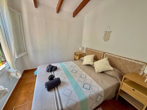 A bedroom with a bed, pillows, a nightstand, a window with curtains, and a small dog. - Garbi: Newly Renovated Apartment with a Pool (S'Arenal D'En Castell (Menorca) (Urbanitzacio))