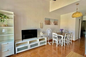 A living room with a flat-screen TV, a white entertainment unit, a dining table with chairs, a fan, and a hanging light fixture. - Garbi: Newly Renovated Apartment with a Pool (S'Arenal D'En Castell (Menorca) (Urbanitzacio))