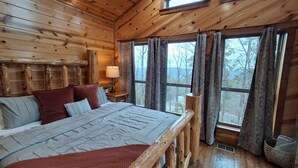 Un dormitorio de cabaña de madera con una cama grande, una mesita de noche y una ventana con cortinas.