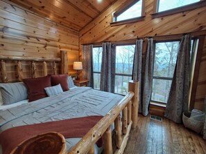 A wooden cabin bedroom with a large bed, a nightstand, and a window with curtains. - Three Bears Cabin | Included Heated Pool, Hot Tub & Mountain Views Near Hochatown | Pet-Friendly (Broken Bow)