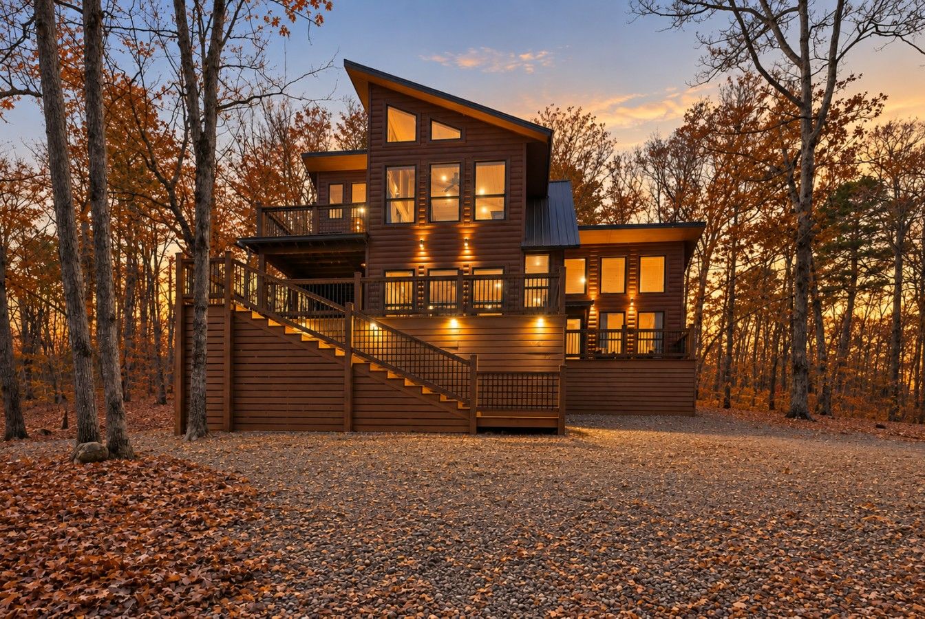 A two-story wooden cabin with a staircase leading to a balcony, surrounded by trees and a gravel driveway.