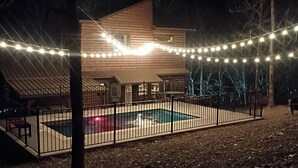 Una casa de madera con piscina cercada, rodeada de árboles y luces colgantes.