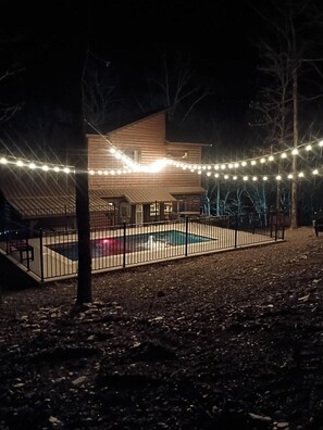 A wooden house with a fenced pool area, surrounded by trees and string lights. - Three Bears Cabin | Included Heated Pool, Hot Tub & Mountain Views Near Hochatown | Pet-Friendly (Broken Bow)
