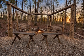 Une pergola en bois avec deux bancs et un foyer central, située dans une zone boisée.
