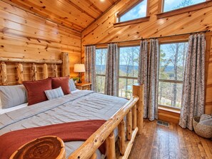 A wooden cabin bedroom with a bed, pillows, curtains, and a window with a view. - Three Bears – Mountainside Cabin with Private Heated Pool & Hot Tub (Broken Bow)
