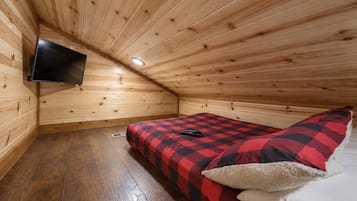 Una habitación pequeña y acogedora con techo inclinado, una cama con manta a cuadros, un televisor montado y piso de madera.