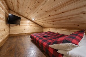 Une petite pièce chaleureuse avec un plafond en pente, un lit recouvert d’une couverture à carreaux, un téléviseur fixé au mur et un plancher en bois.