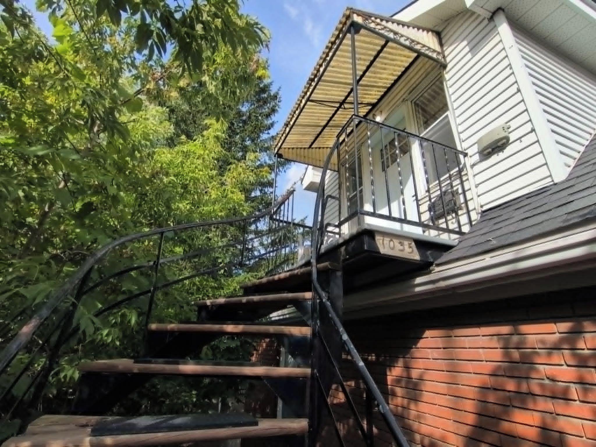 Un escalier en bois mène à un balcon doté d’une rambarde en métal, qui est adjacent à un mur de briques et à un bâtiment blanc avec une fenêtre.