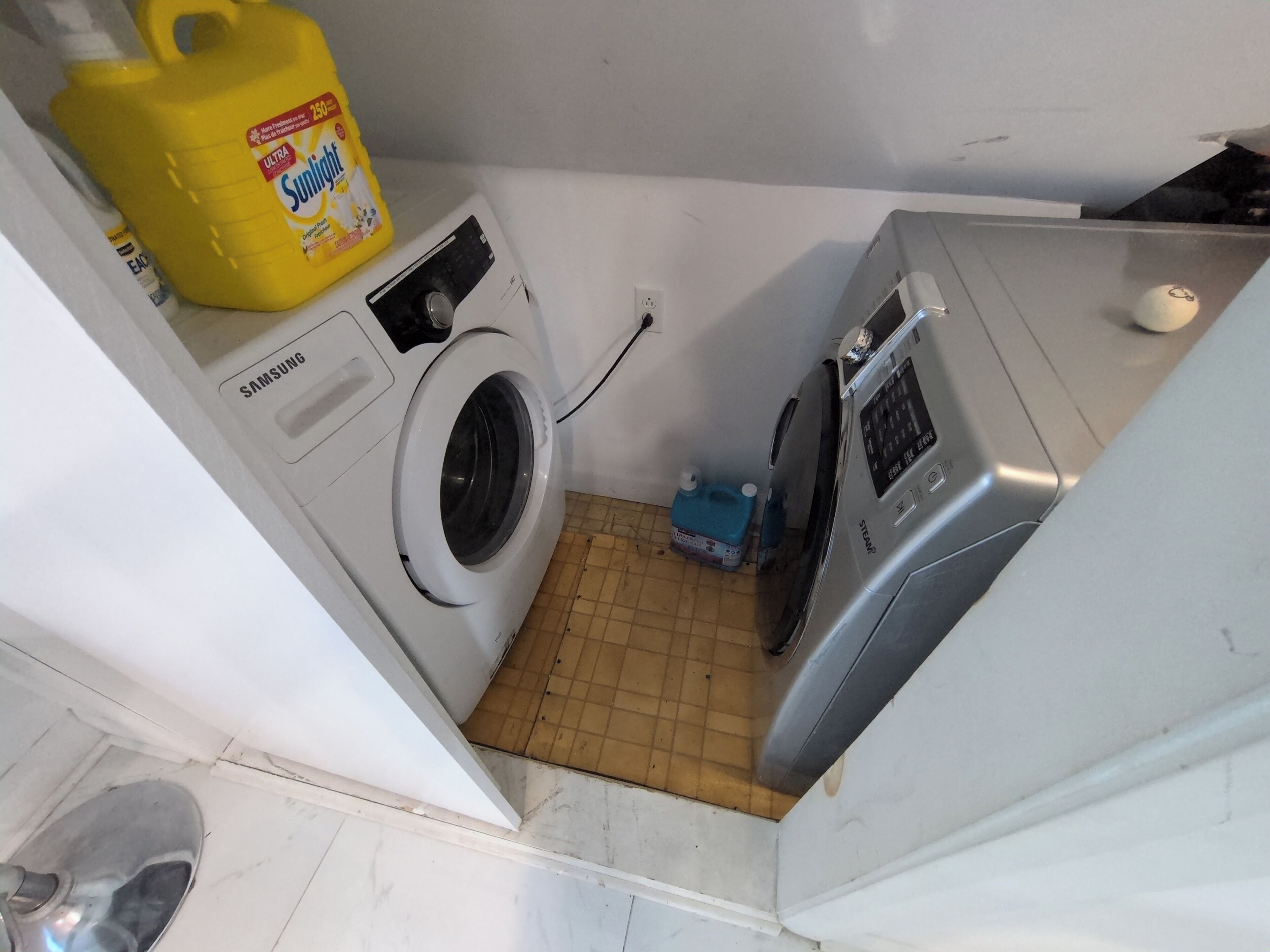 Un espace buanderie compact comprenant un lave-linge et un sèche-linge Samsung, un récipient jaune pour la lessive et un bidon bleu pour l’eau.