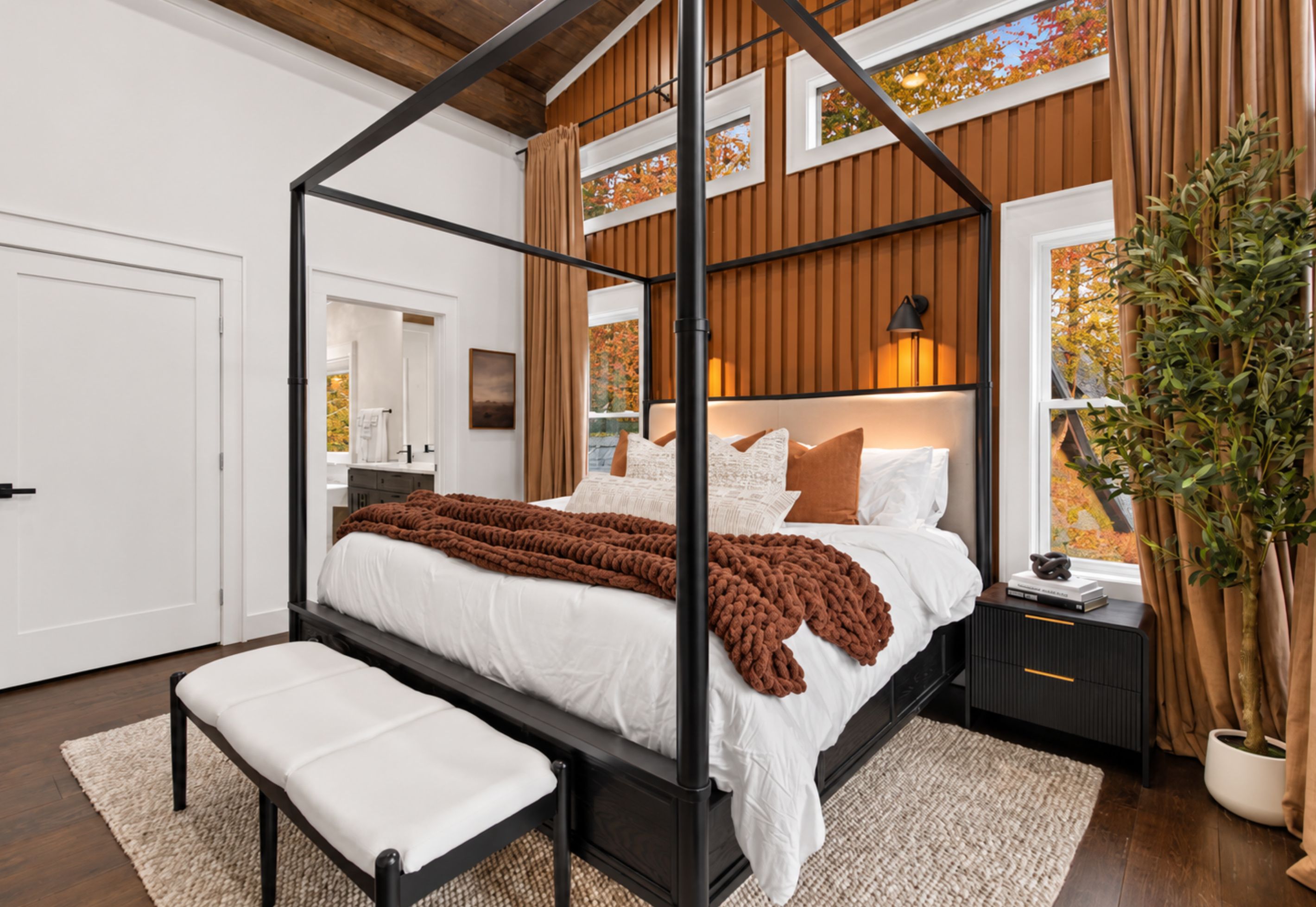 A four-poster bed with a brown throw blanket, a bedside table with a lamp, and a potted plant.