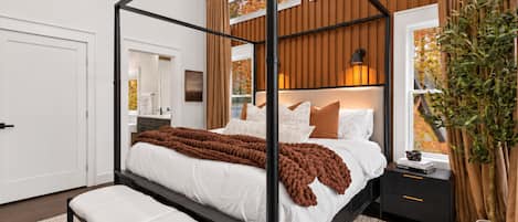 A four-poster bed with a brown throw blanket, a bedside table with a lamp, and a potted plant.