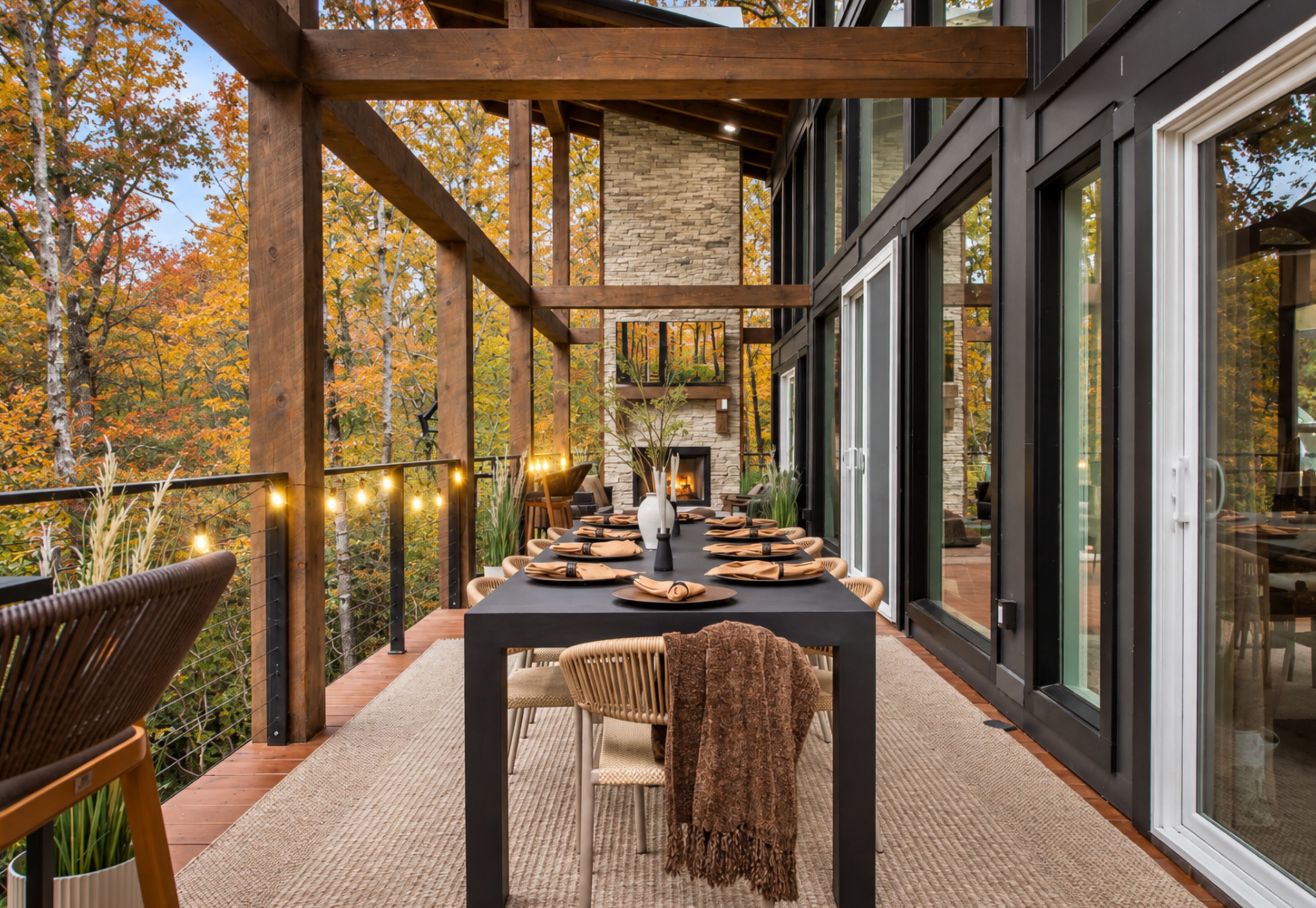 A covered outdoor dining area with a stone fireplace, wooden beams, and a view of autumn trees.
