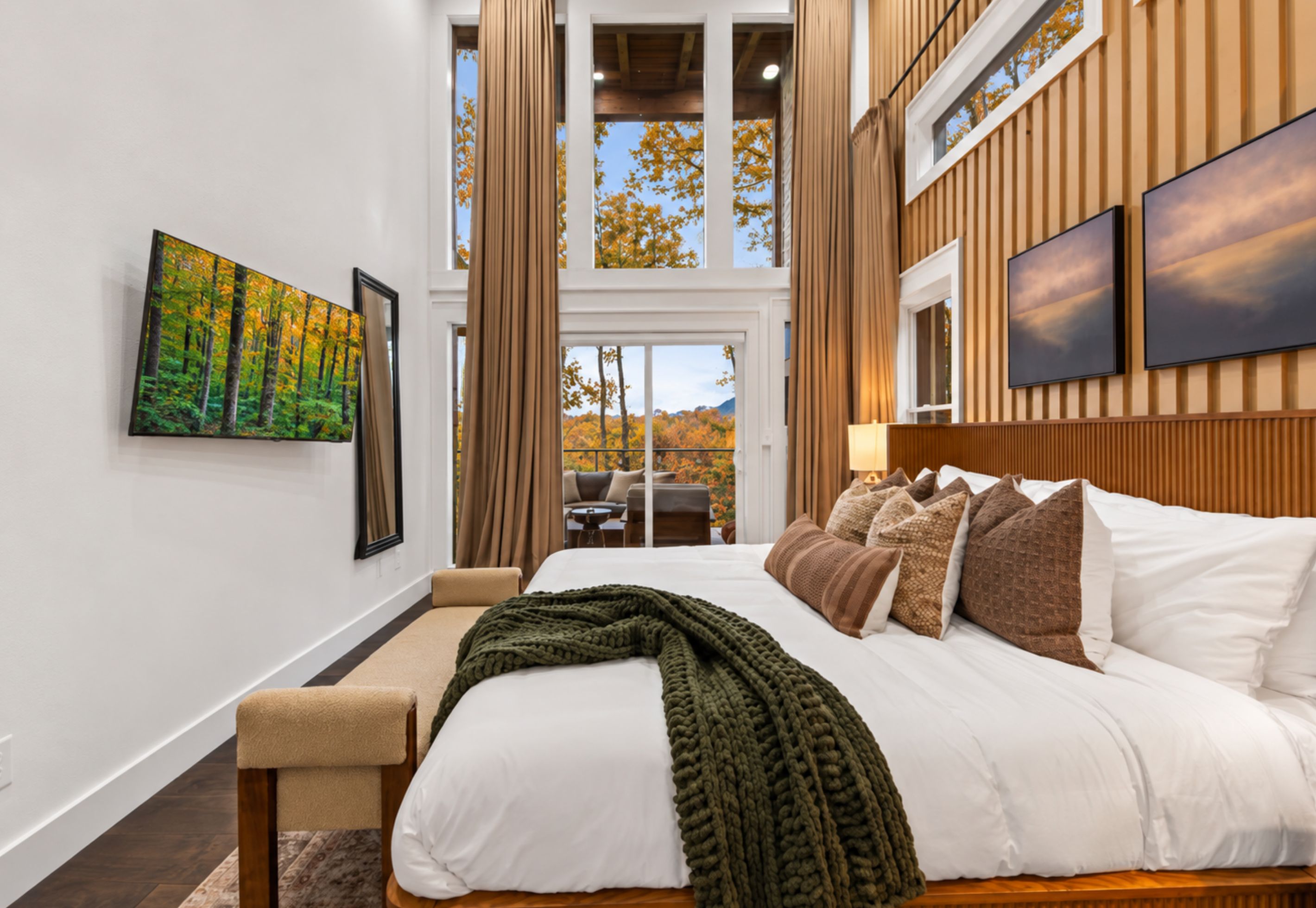 A modern hotel room with a large bed, a view of autumn trees, and a painting of a forest.