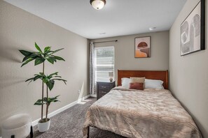 Een slaapkamer met een bed, een nachtkastje, een kamerplant, een raam met jaloezieën en een plafondlamp.
