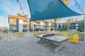 Un espace extérieur aménagé en terrasse, avec une table et des chaises, une grande voile d’ombrage et une rangée de bâtiments en arrière-plan.