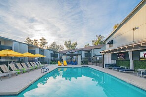 Ein Swimmingpool mit Liegestühlen und Sonnenschirmen, umgeben von Gebäuden mit Balkonen und einer überdachten Sitzgelegenheit.