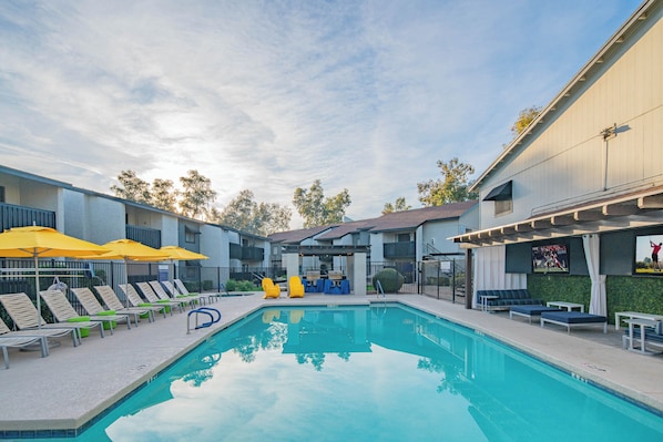 A swimming pool with lounge chairs and umbrellas, surrounded by buildings with balconies and a covered seating area.