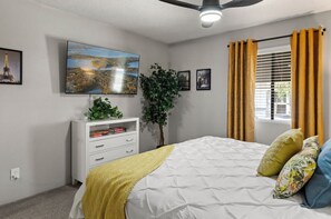 Ein Schlafzimmer mit einem Bett, einem an der Wand befestigten Fernseher, einer Pflanze und einem Fenster mit Vorhängen.