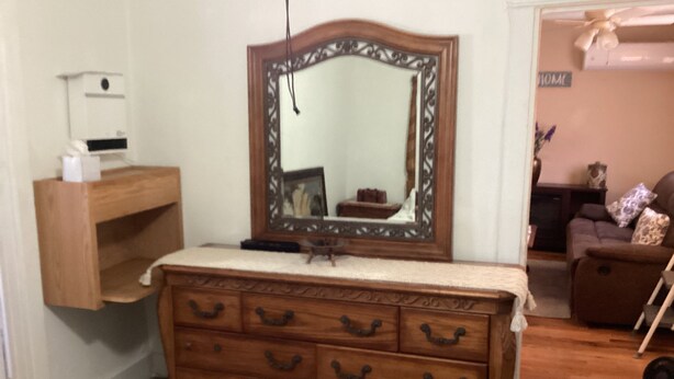 Une commode en bois avec un miroir au-dessus, une petite étagère et, en arrière-plan, un panneau indiquant le mot « MAISON ».