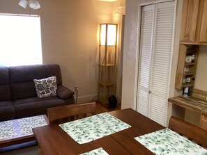 A living room with a brown sofa, a wooden dining table with floral placemats, a floor lamp, and a closed white door. - Cozy 1-bedroom apartment in beautiful Albuquerque with AC private gated parking (Albuquerque)