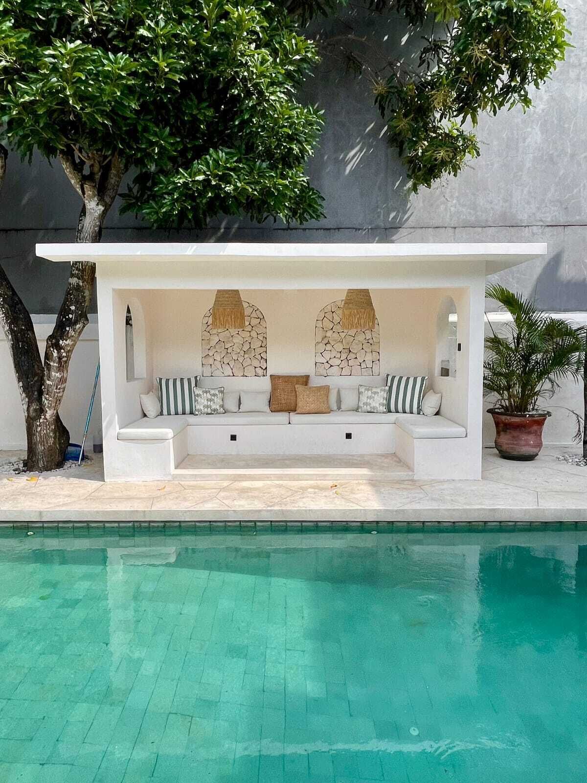 Área de estar junto à piscina, com estrutura branca, detalhes em parede de pedra e planta em vaso.