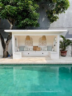 Un área de descanso junto a la piscina con una estructura blanca, detalles de pared de piedra y una planta en maceta.