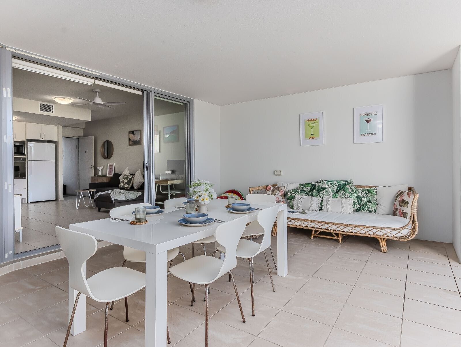 A modern dining area with a white table and chairs, a sofa with a tropical print, and a kitchen in the background.