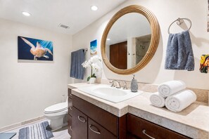 Une salle de bain avec un grand miroir ovale, un lavabo blanc et des toilettes. On y trouve des serviettes et une petite plante sur le comptoir.