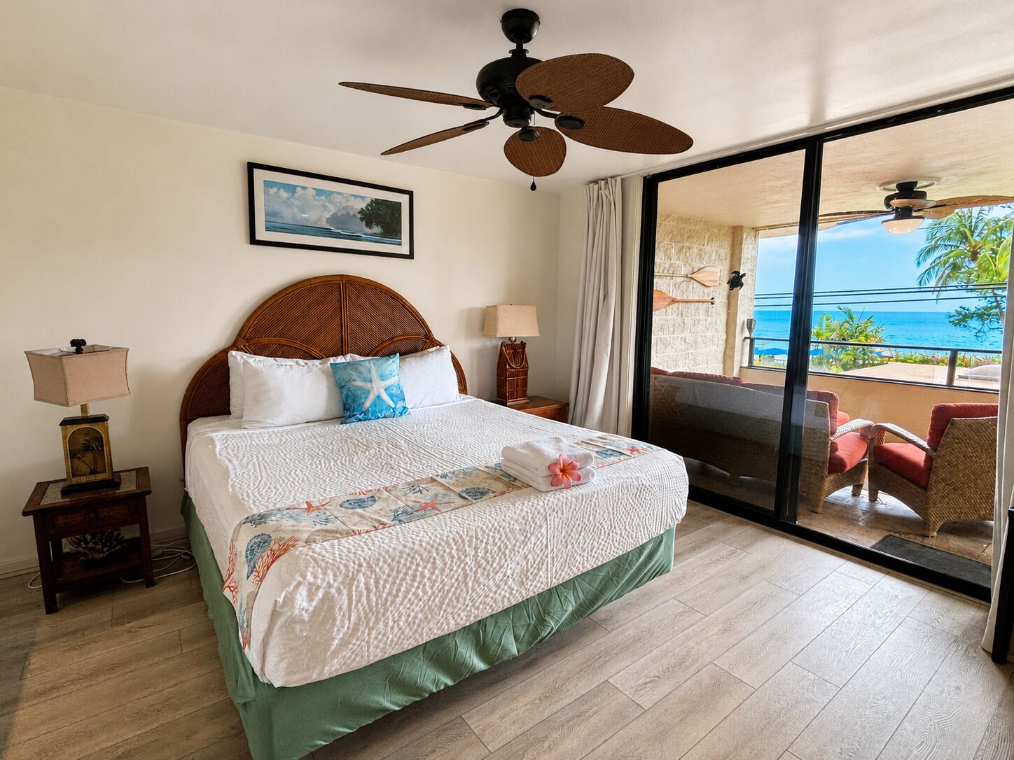 Habitación de hotel con cama, ventilador de techo, ventana con vista al océano y balcón con sillas.