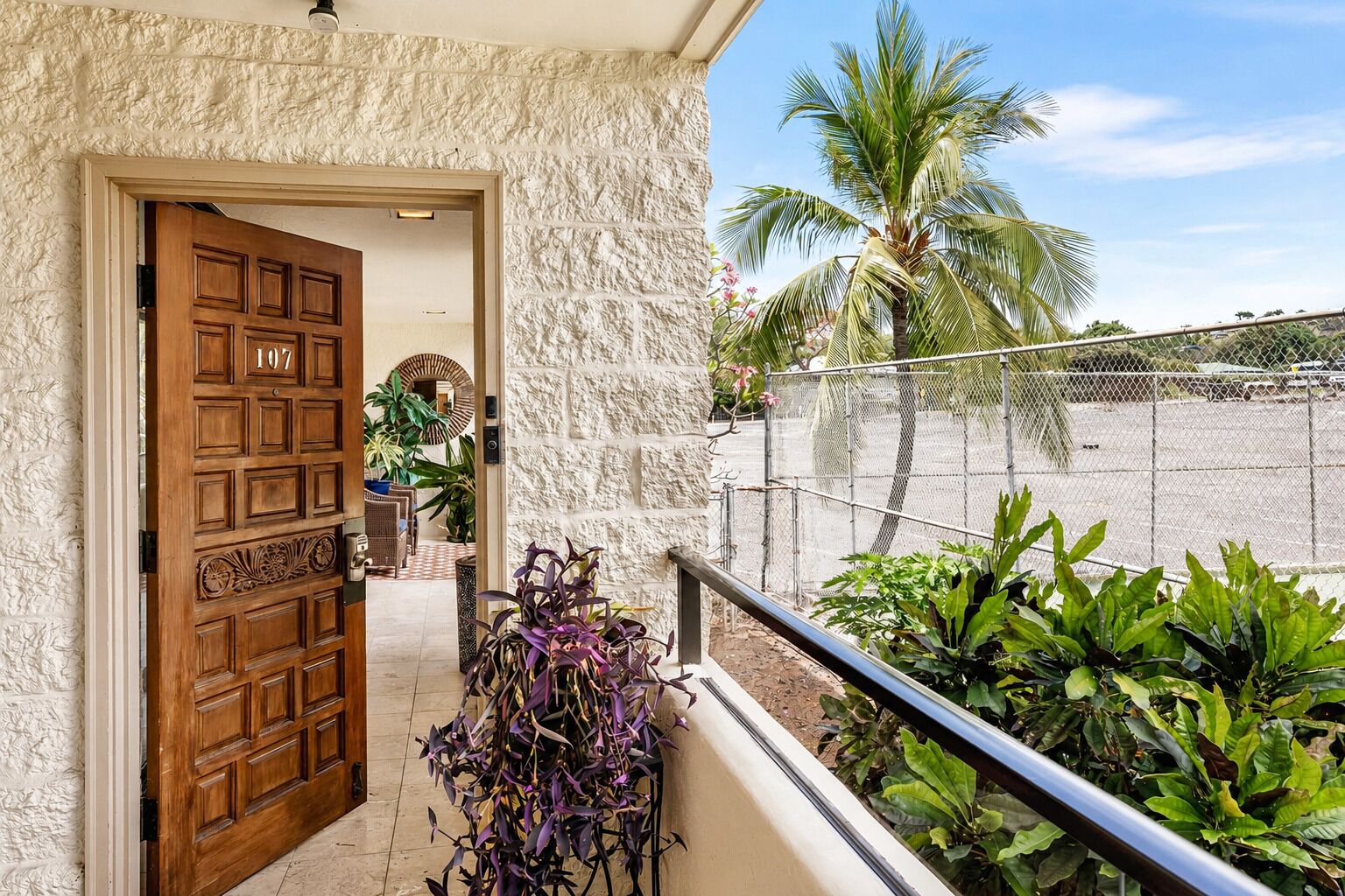 Entrada de hotel con puerta de madera marcada '107', pared de piedra y vista de una palmera y una cerca.
