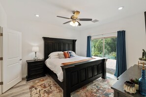 A bedroom with a dark wooden bed, a ceiling fan, a nightstand with a lamp, and a view of greenery through a large window. - Peaceful 3BR Getaway | Pines, Wildlife, Hiking & Golf (Payson)