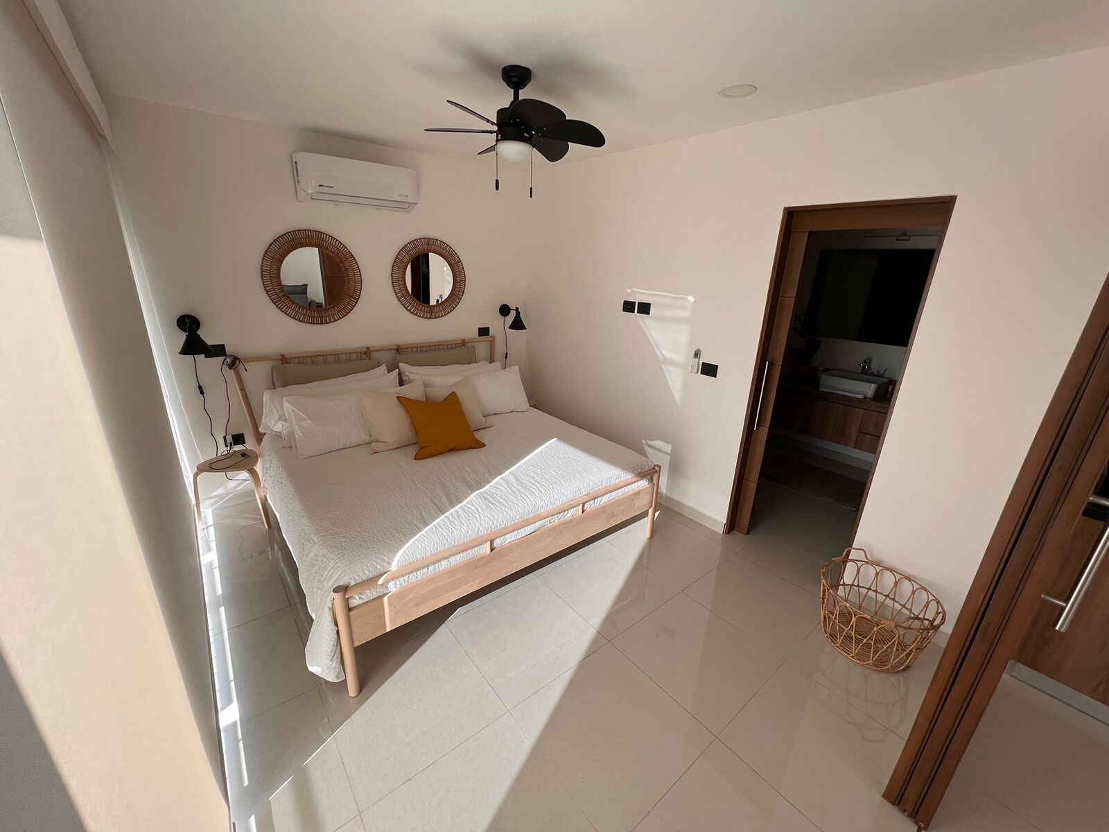 A bedroom with a bed, a ceiling fan, two wall-mounted mirrors, an air conditioning unit, and a doorway leading to a bathroom.