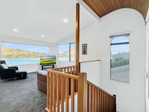 A modern living room with a large flat-screen TV, a wooden staircase, and a view of a body of water through large windows. - Lakeview Townhouse | Relax Above Queenstown (Queenstown)