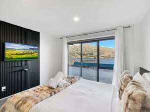A hotel room with a bed, a television mounted on the wall, and a large window offering a view of a mountainous landscape. - Stunning Lake Views | Sunny Corner Townhouse (Queenstown)