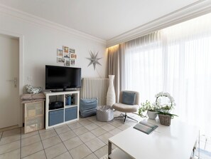 Un salon moderne doté d’un téléviseur à écran plat, d’une table basse blanche, d’un fauteuil confortable et d’une grande fenêtre ornée de rideaux légers.