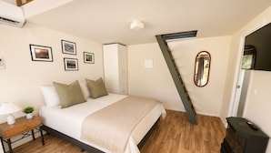 A bedroom with a bed, a desk with a lamp, a potted plant, a mirror, and a black stove. - Tasman Holiday Parks Mount Beauty (Tawonga South)
