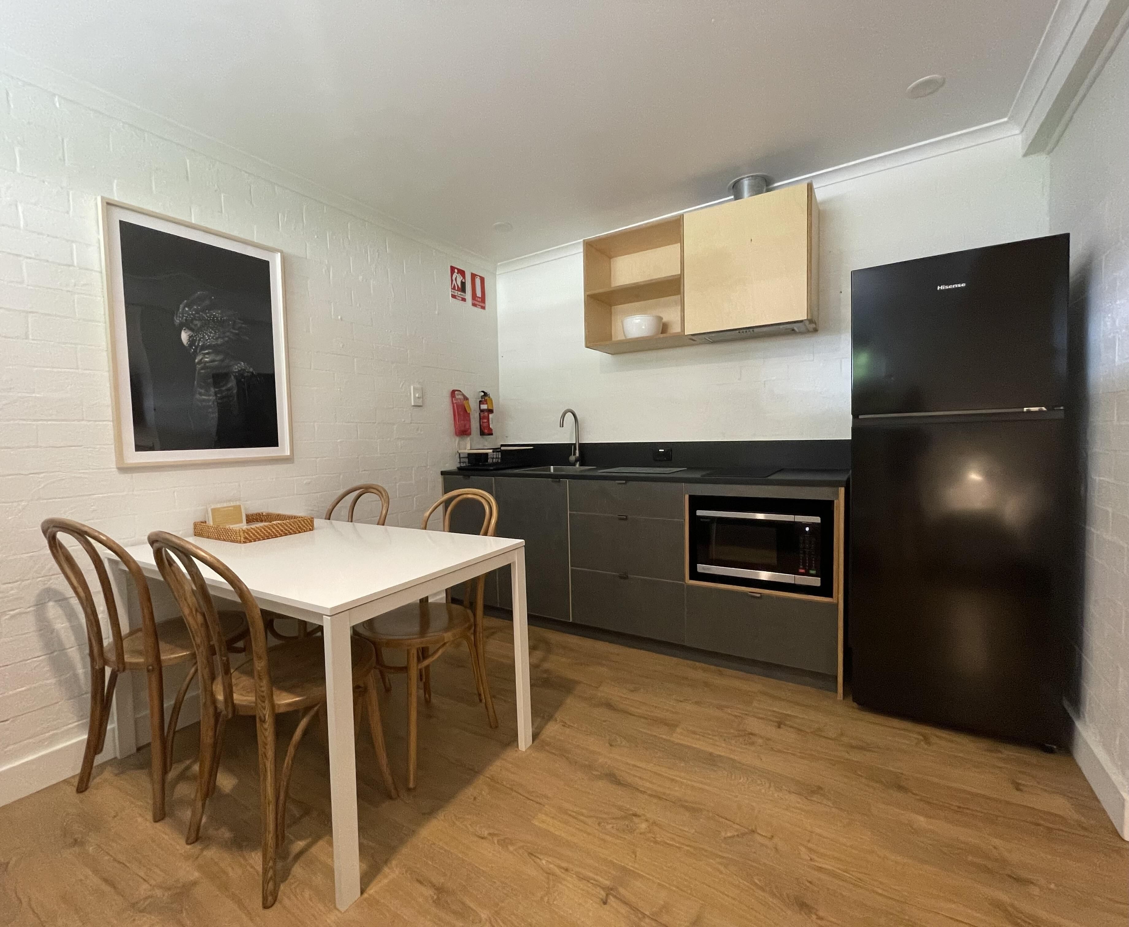 Une cuisine compacte comprenant une table à manger, des chaises, un réfrigérateur noir, un four à micro-ondes et une étagère en bois au-dessus de l’évier.