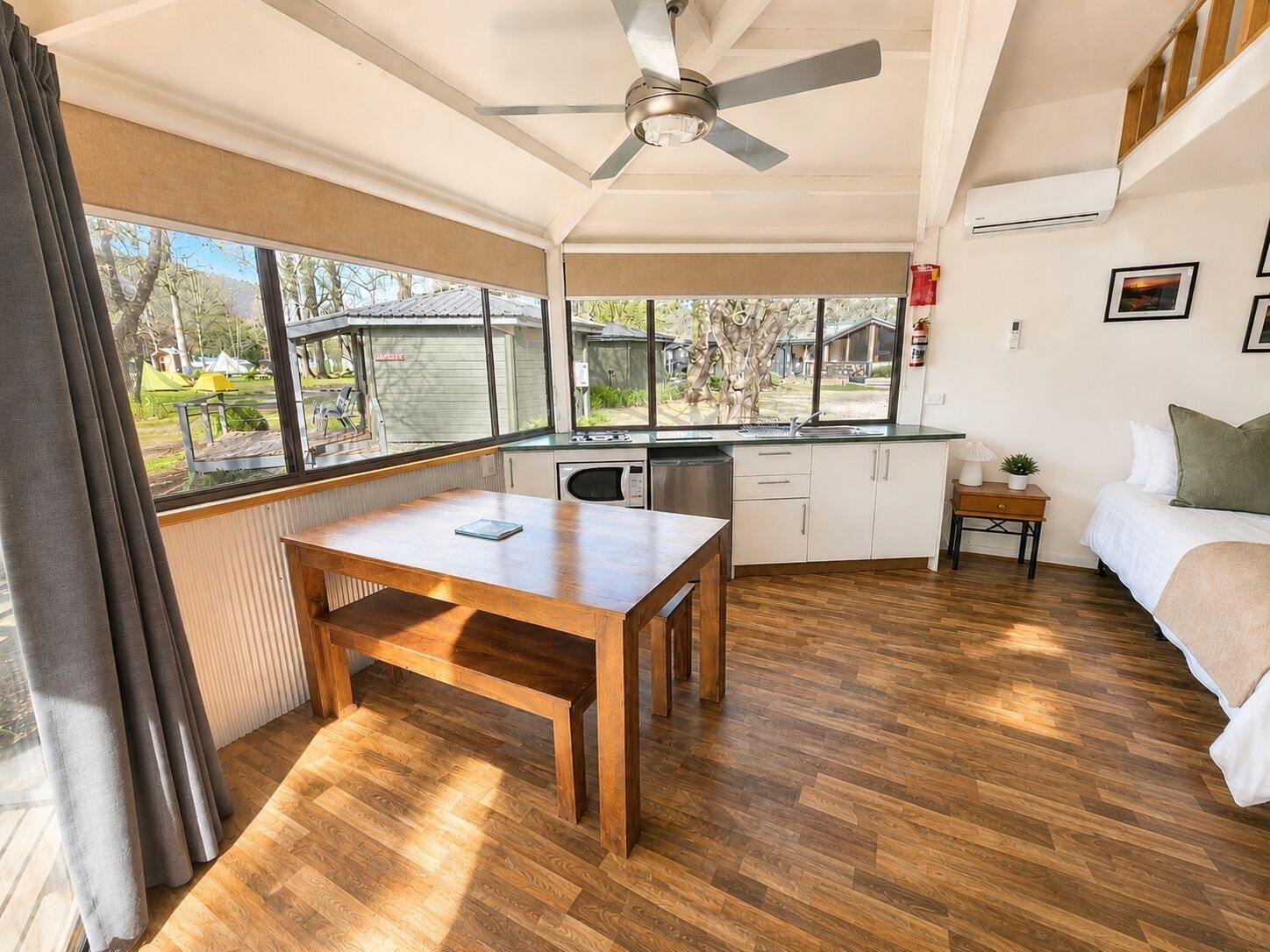 A compact room with a wooden floor, a dining table, a bed, a kitchenette, and large windows offering a view of the outdoors.