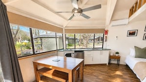 A compact room with a wooden floor, a dining table, a bed, a kitchenette, and large windows offering a view of the outdoors. - Tasman Holiday Parks Mount Beauty (Tawonga South)