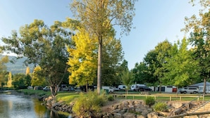 A lakeside camping area with RVs parked, a picnic table, and a wooden bridge over the water. - Tasman Holiday Parks Mount Beauty (Tawonga South)