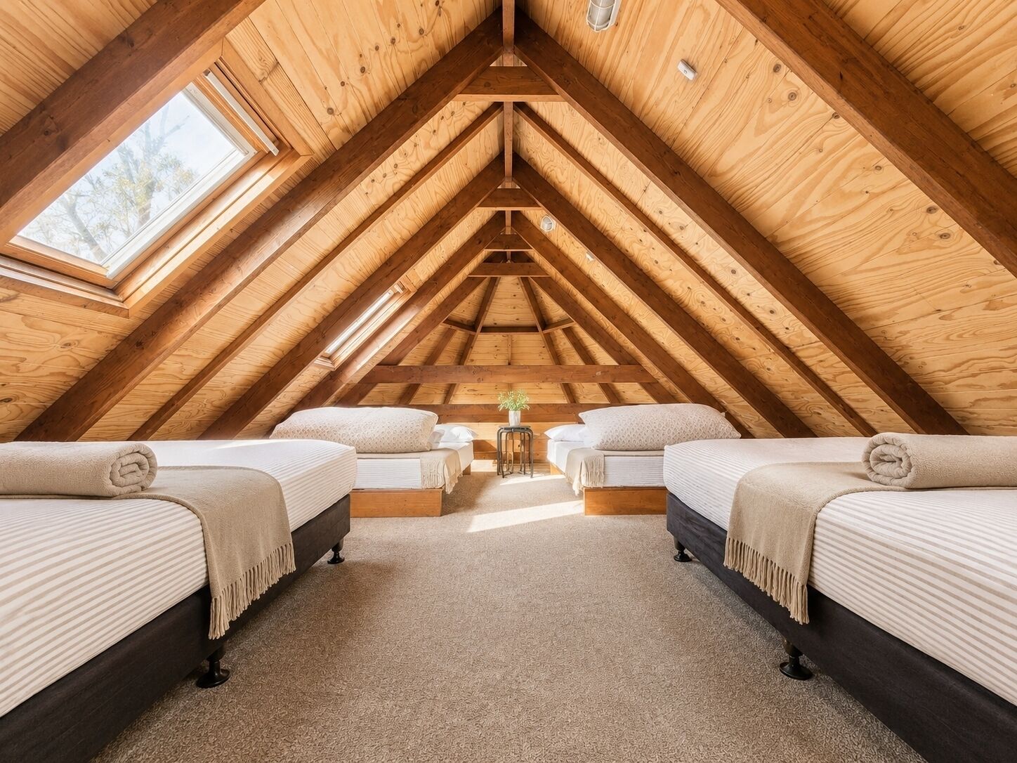 A room with a sloped ceiling, wooden beams, two beds with headboards, a small table, and a window.