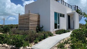Un edificio moderno de dos pisos con una rampa de hormigón que conduce a la entrada, rodeado de vegetación y un cielo despejado.