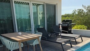 Zona junto a la piscina con mesa y sillas, una parrilla y vistas al entorno verde.